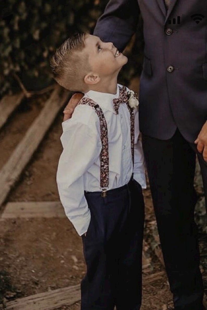 May include: A young boy in a white button-down shirt, navy pants, and floral suspenders and bow tie. He is looking up with a smile. The boy is standing next to a person wearing a dark suit.