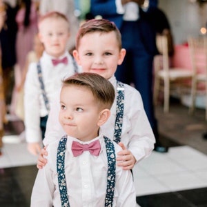 Puede incluir: Tres ni&ntilde;os peque&ntilde;os vestidos con camisas blancas, pajaritas a juego y tirantes florales. Los ni&ntilde;os sonr&iacute;en. La imagen est&aacute; tomada en una boda o evento especial.