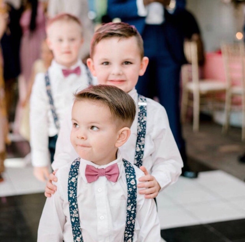 Puede incluir: Tres ni&ntilde;os peque&ntilde;os vestidos con camisas blancas, pajaritas a juego y tirantes florales. Los ni&ntilde;os sonr&iacute;en. La imagen est&aacute; tomada en una boda o evento especial.