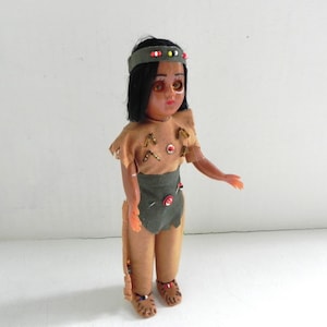 Può includere: Una bambola vintage con capelli scuri e occhi marroni, vestita con un abito marrone e verde con accenti di perline. La bambola è vestita come una nativa americana.