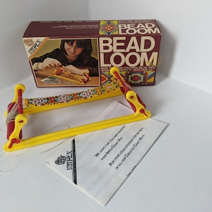 Può includere: Un telaio per perline in plastica giallo e rosso con un progetto di perline parzialmente completato. Il telaio è su una superficie bianca con un libretto di istruzioni bianco e nero. La scatola del telaio per perline è sullo sfondo. La scatola è rossa e marrone con le parole "Bead Loom" in bianco.