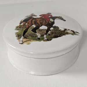 Może przedstawiać: Biała ceramiczna szkatułka z okrągłą pokrywką przedstawiającą vintageową ilustrację konia i jeźdźca w scenie polowania.