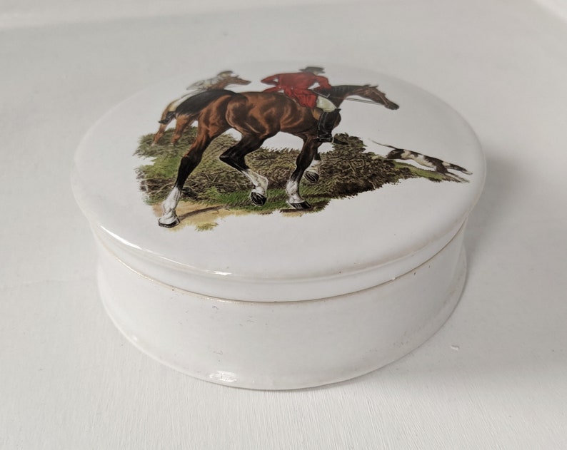 Może przedstawiać: Biała ceramiczna szkatułka z okrągłą pokrywką przedstawiającą vintageową ilustrację konia i jeźdźca w scenie polowania.