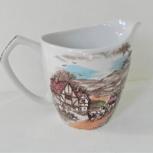 May include: White ceramic pitcher with a brown and green countryside scene featuring a house, trees, and people riding in a horse-drawn carriage.
