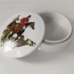 Może przedstawiać: Białe ceramiczne pudełko na drobiazgi z pokrywką przedstawiającą kolorową ilustrację dw&oacute;ch os&oacute;b na koniach jadących przez pole. Pokrywka jest okrągła, a pudełko jest okrągłe z lekko uniesionym rantem.