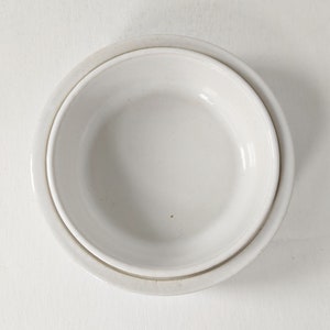 Może przedstawiać: Biała ceramiczna miska włożona w nieco większą białą ceramiczną miskę. Obie miski są gładkie i mają lekko błyszczące wykończenie.
