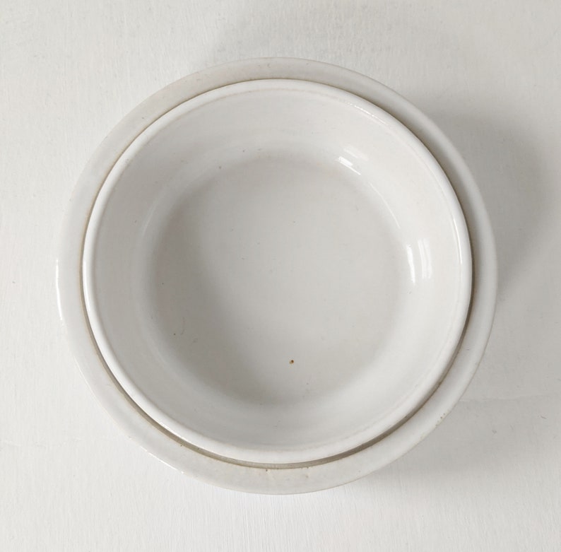 Może przedstawiać: Biała ceramiczna miska włożona w nieco większą białą ceramiczną miskę. Obie miski są gładkie i mają lekko błyszczące wykończenie.