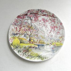 Può includere: Un piatto in ceramica bianca con una scena dipinta di un cottage incastonato in un lussureggiante paesaggio verde con un fiume che lo attraversa. Il cottage ha un tetto di paglia e una piccola finestra. Gli alberi sono in fiore con fiori rosa e bianchi.