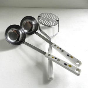 May include: Three kitchen utensils with white handles and black and gold diamond patterns. The utensils are a potato masher, a ladle, and a smaller ladle.