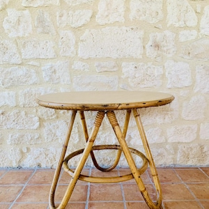 May include: A round, light brown bamboo table with a woven design. The table has a light brown top and four legs that curve outwards.