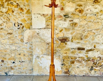 Può includere: Un attaccapanni in legno color marrone chiaro con un'asta centrale verticale, una sezione superiore a più bracci con ganci curvi per appendere indumenti e una base a tre piedi rinforzata da un anello di supporto circolare alla base.