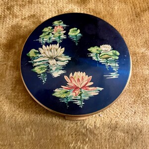 Op de afbeelding: Een ronde, marineblauwe compact met een goudkleurige rand. De compact heeft een geschilderd ontwerp van witte en roze waterlelies met groene leliebladeren die op een reflecterend wateroppervlak drijven.