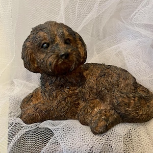 Puede incluir: Figura detallada de un perro pequeño, tumbado con las patas delanteras extendidas. El perro tiene pelaje marrón y negro, con un aspecto texturizado. El fondo es una tela suave y blanca.