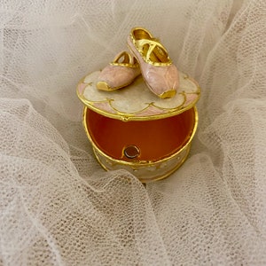 Puede incluir: Una caja de joyería decorativa con tapa con bisagras, con un par de zapatillas de ballet rosas. La caja es redonda, con un borde dorado y un diseño floral rosa y blanco. El interior es de color melocotón.