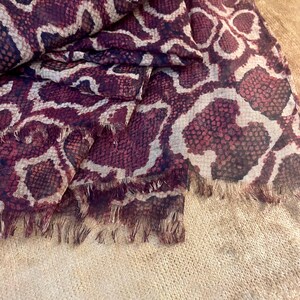 Puede incluir: Bufanda burdeos y crema con estampado de piel de serpiente. La bufanda tiene un borde con flecos y está drapeada sobre una superficie beige. El patrón presenta un diseño repetitivo de formas burdeos oscuras y crema.