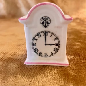 Könnte beinhalten: Eine kleine, weiße Keramikuhr mit rosa Zierleiste. Das Zifferblatt hat schwarze römische Ziffern und Zeiger, die auf 3 Uhr zeigen. Über dem Zifferblatt befindet sich ein dekoratives schwarzes Design. Die Uhr ist ein dekorativer Miniaturartikel.