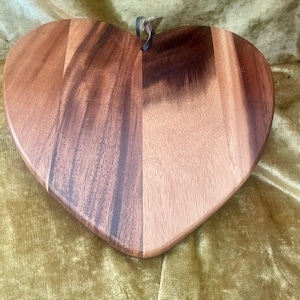 May include: Heart-shaped wooden cutting board with a natural wood grain finish. The board is made of multiple wood panels and has a leather loop for hanging. The wood tones range from light to dark brown.