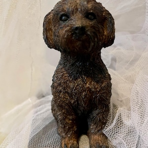 May include: A detailed, brown dog figurine with a textured coat and dark eyes. The dog is sitting upright, with its front paws visible. The figurine has a realistic appearance and is set against a soft, white background.