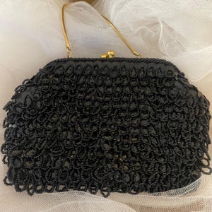 May include: A vintage black beaded handbag with a gold-tone handle. The bag features a detailed, textured pattern of small beads, creating a decorative surface. The clasp is gold-toned. The bag is set against a soft, white background.