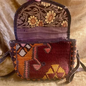May include: Open, vintage shoulder bag with a tapestry design in shades of burgundy, orange, and yellow. The interior flap features a floral pattern with cream and yellow flowers on a brown background. The bag has a dark brown leather strap.