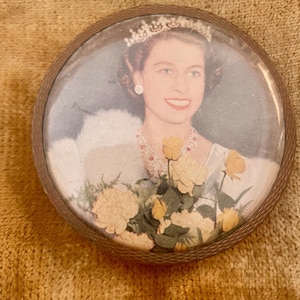 Può includere: Un portacipria rotondo vintage con un ritratto di donna con tiara, orecchini e collana. Il ritratto è racchiuso in una cornice color bronzo e presenta un bouquet di fiori gialli.
