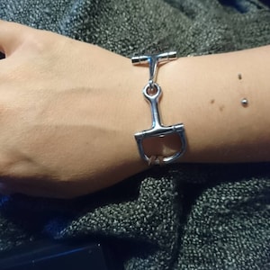 May include: A silver bracelet with a snaffle bit design. The bracelet is on a person's wrist.