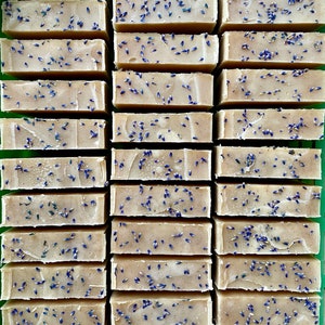 Puede incluir: Una colecci&oacute;n de pastillas de jab&oacute;n rectangulares de color beige dispuestas en filas. Cada pastilla presenta una superficie lisa de color tostado claro con peque&ntilde;os capullos de lavanda morados secos esparcidos por encima.