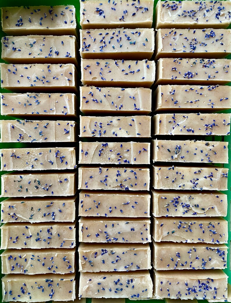 Puede incluir: Una colecci&oacute;n de pastillas de jab&oacute;n rectangulares de color beige dispuestas en filas. Cada pastilla presenta una superficie lisa de color tostado claro con peque&ntilde;os capullos de lavanda morados secos esparcidos por encima.
