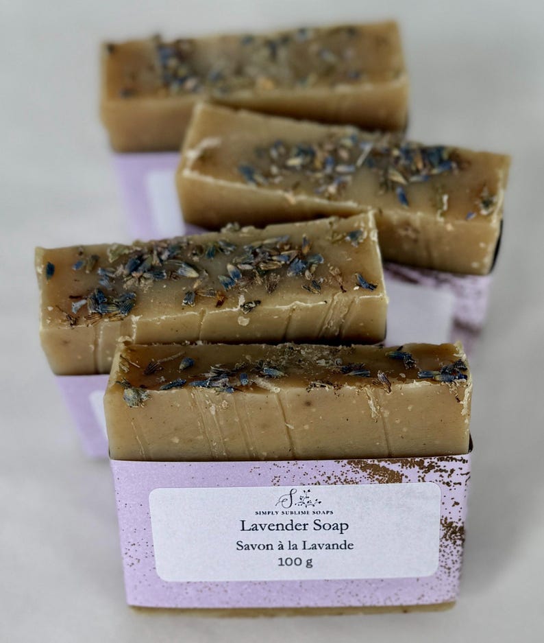 Puede incluir: Cuatro barras de jab&oacute;n rectangulares de color beige cubiertas con brotes de lavanda secos. Cada barra est&aacute; envuelta en una banda de papel de color violeta claro con el texto Simply Sublime Soaps, Lavender Soap, Savon &agrave; la Lavande, 100 g.