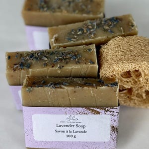 Puede incluir: Barras de jab&oacute;n rectangulares de color beige con brotes de lavanda secos, envueltas en una etiqueta de papel morado claro con el texto Simply Sublime Soaps, Lavender Soap, Savon &agrave; la Lavande, 100 g. Una esponja vegetal de lufa beige y fibrosa.
