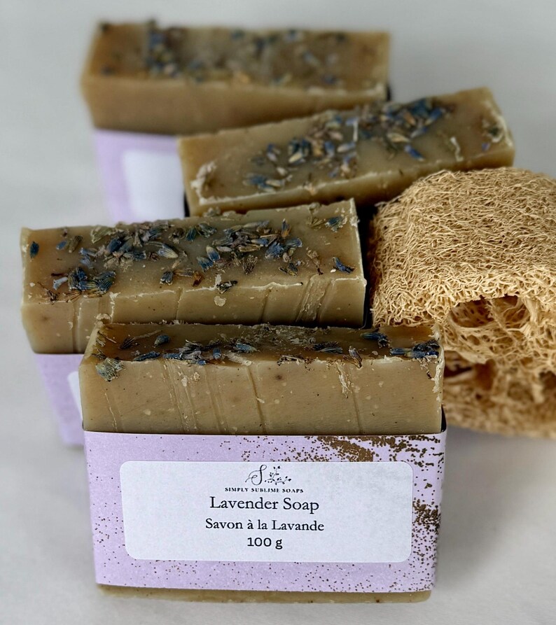 Puede incluir: Barras de jab&oacute;n rectangulares de color beige con brotes de lavanda secos, envueltas en una etiqueta de papel morado claro con el texto Simply Sublime Soaps, Lavender Soap, Savon &agrave; la Lavande, 100 g. Una esponja vegetal de lufa beige y fibrosa.