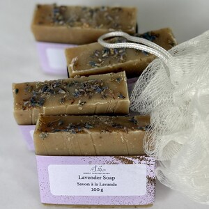 Puede incluir: Cuatro barras rectangulares de jab&oacute;n de lavanda color canela cubiertas con capullos de flores moradas secas. Cada barra est&aacute; envuelta en una etiqueta de papel morado claro con el texto Simply Sublime Soaps, Lavender Soap, Savon &agrave; la Lavande y 100 g. Una esponja de ba&ntilde;o de malla blanca.