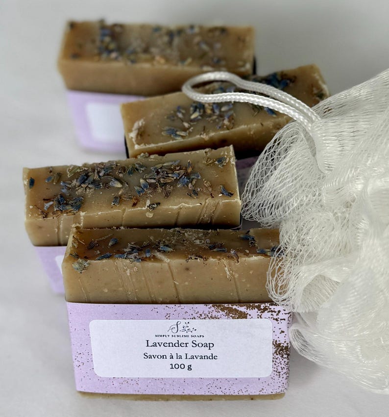 Puede incluir: Cuatro barras rectangulares de jab&oacute;n de lavanda color canela cubiertas con capullos de flores moradas secas. Cada barra est&aacute; envuelta en una etiqueta de papel morado claro con el texto Simply Sublime Soaps, Lavender Soap, Savon &agrave; la Lavande y 100 g. Una esponja de ba&ntilde;o de malla blanca.