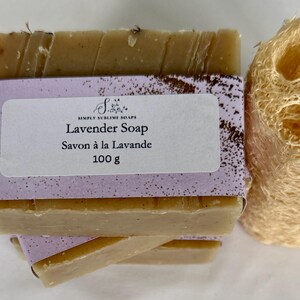 Puede incluir: Una pastilla de jab&oacute;n rectangular de color beige envuelta en una etiqueta de papel morado claro con el texto Simply Sublime Soaps, Lavender Soap, Savon &agrave; la Lavande, 100 g. Una esponja de lufa cil&iacute;ndrica beige se encuentra junto al jab&oacute;n.