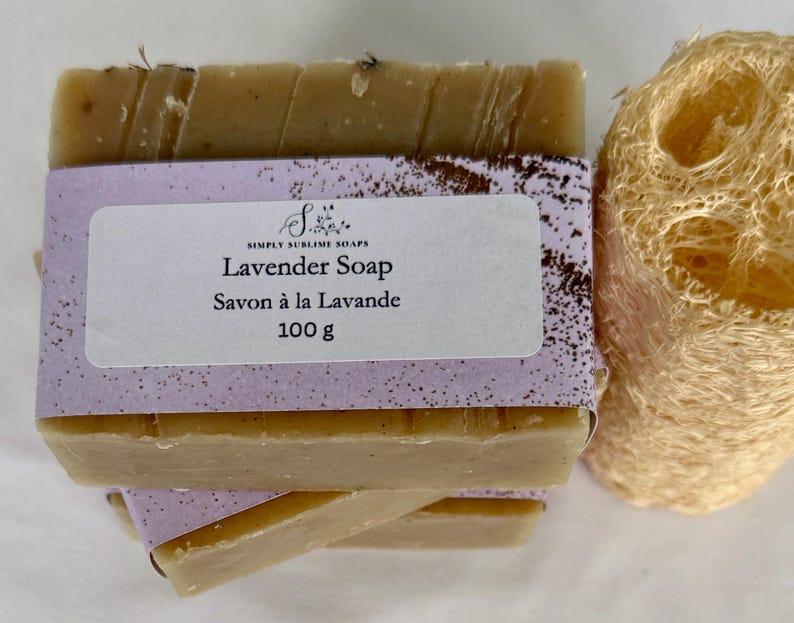 Puede incluir: Una pastilla de jab&oacute;n rectangular de color beige envuelta en una etiqueta de papel morado claro con el texto Simply Sublime Soaps, Lavender Soap, Savon &agrave; la Lavande, 100 g. Una esponja de lufa cil&iacute;ndrica beige se encuentra junto al jab&oacute;n.