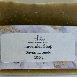 Puede incluir: Una pastilla de jab&oacute;n rectangular de tono oliva envuelta en una banda de papel con un patr&oacute;n de m&aacute;rmol morado y dorado. La etiqueta muestra el texto Simply Sublime Soaps, Lavender Soap, Savon Lavande y el peso de 100 g en letras negras.