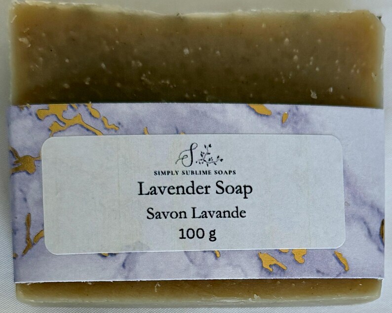 Puede incluir: Una pastilla de jab&oacute;n rectangular de tono oliva envuelta en una banda de papel con un patr&oacute;n de m&aacute;rmol morado y dorado. La etiqueta muestra el texto Simply Sublime Soaps, Lavender Soap, Savon Lavande y el peso de 100 g en letras negras.
