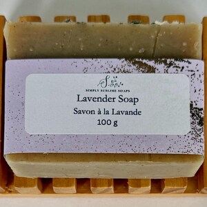 Puede incluir: Una pastilla de jab&oacute;n rectangular de color beige con aroma a lavanda sobre una jabonera de madera con listones. El jab&oacute;n est&aacute; envuelto en una banda de papel morado claro con el texto Simply Sublime Soaps, Lavender Soap, Savon &agrave; la Lavande y 100 g.