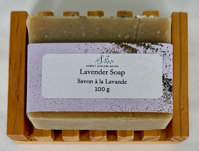 Puede incluir: Una pastilla de jab&oacute;n rectangular de color beige con aroma a lavanda sobre una jabonera de madera con listones. El jab&oacute;n est&aacute; envuelto en una banda de papel morado claro con el texto Simply Sublime Soaps, Lavender Soap, Savon &agrave; la Lavande y 100 g.