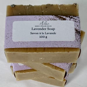Puede incluir: Una pila de barras rectangulares de jab&oacute;n de lavanda beige envueltas en una banda de papel de color violeta claro. La etiqueta presenta el texto Simply Sublime Soaps, Lavender Soap, Savon &agrave; la Lavande y 100 g. El jab&oacute;n tiene una textura suave con motas oscuras visibles.
