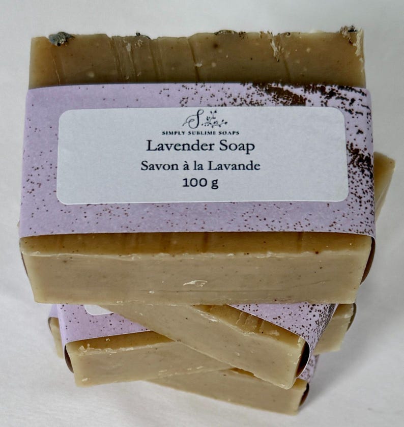 Puede incluir: Una pila de barras rectangulares de jab&oacute;n de lavanda beige envueltas en una banda de papel de color violeta claro. La etiqueta presenta el texto Simply Sublime Soaps, Lavender Soap, Savon &agrave; la Lavande y 100 g. El jab&oacute;n tiene una textura suave con motas oscuras visibles.