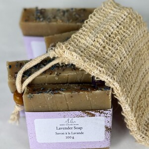 Puede incluir: Pastillas de jab&oacute;n rectangulares de color marr&oacute;n con capullos de flores secas, envueltas en una etiqueta de papel morado claro con el texto Simply Sublime Soaps Lavender Soap Savon &agrave; la Lavande 100 g, junto a una bolsa de sisal beige.