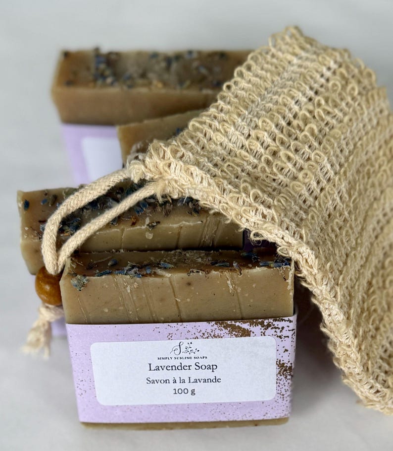 Puede incluir: Pastillas de jab&oacute;n rectangulares de color marr&oacute;n con capullos de flores secas, envueltas en una etiqueta de papel morado claro con el texto Simply Sublime Soaps Lavender Soap Savon &agrave; la Lavande 100 g, junto a una bolsa de sisal beige.