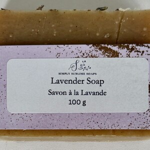 Puede incluir: Una pastilla de jab&oacute;n rectangular de color tostado envuelta en una banda de papel morado claro. La etiqueta muestra el texto Simply Sublime Soaps, Lavender Soap, Savon &agrave; la Lavande y 100 g. Peque&ntilde;os restos bot&aacute;nicos se ven en el borde superior.