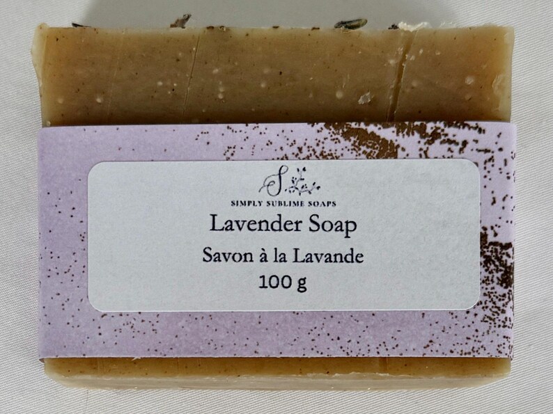 Puede incluir: Una pastilla de jab&oacute;n rectangular de color tostado envuelta en una banda de papel morado claro. La etiqueta muestra el texto Simply Sublime Soaps, Lavender Soap, Savon &agrave; la Lavande y 100 g. Peque&ntilde;os restos bot&aacute;nicos se ven en el borde superior.
