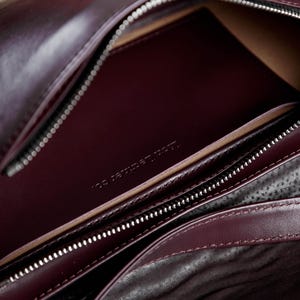 Op de afbeelding: Een diepbordeauxrode leren tas met een gladde buitenkant en een hoofdvak met ritssluiting. De binnenkant toont een bijpassend bordeauxrood leren vak met de in reli&euml;f aangebrachte tekst 'ION LEATHER CO.' en een donkere, gestructureerde voering.