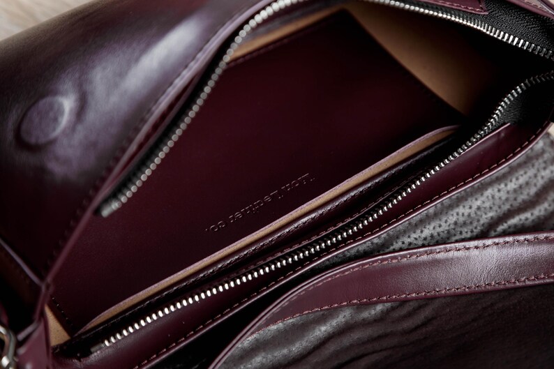 Op de afbeelding: Een diepbordeauxrode leren tas met een gladde buitenkant en een hoofdvak met ritssluiting. De binnenkant toont een bijpassend bordeauxrood leren vak met de in reli&euml;f aangebrachte tekst 'ION LEATHER CO.' en een donkere, gestructureerde voering.