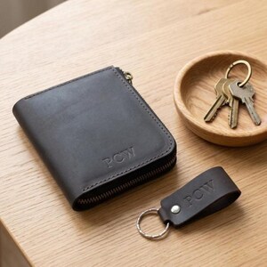 May include: A dark brown leather wallet with a zip closure and the embossed letters PCW, accompanied by a matching leather keyring with the letters PCW and a silver metal ring, plus a set of three silver metal keys on a ring.