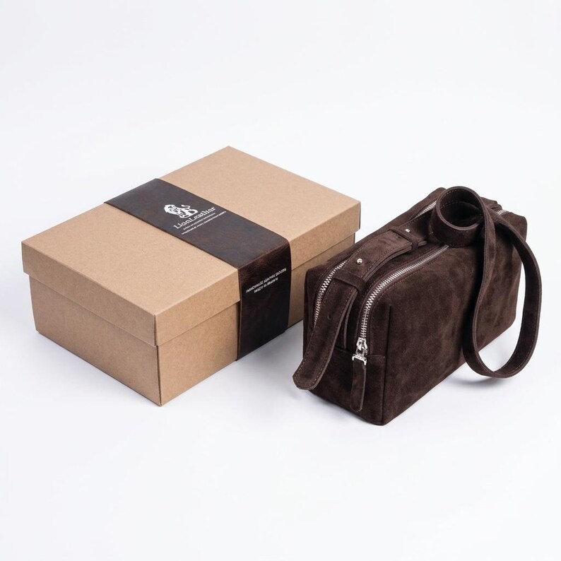 Pu&ograve; includere: Una trousse rettangolare in camoscio marrone scuro con cerniere in metallo argentato e manico abbinato, posizionata accanto a una scatola regalo in cartone con una fascia di carta marrone scuro recante il testo LionLeather.