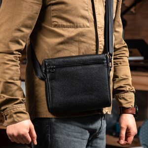 May include: A black pebbled leather crossbody bag featuring a rectangular shape, a top zipper closure, and a wide black adjustable fabric shoulder strap. The bag is worn across the torso, showcasing a textured surface and a minimalist design.